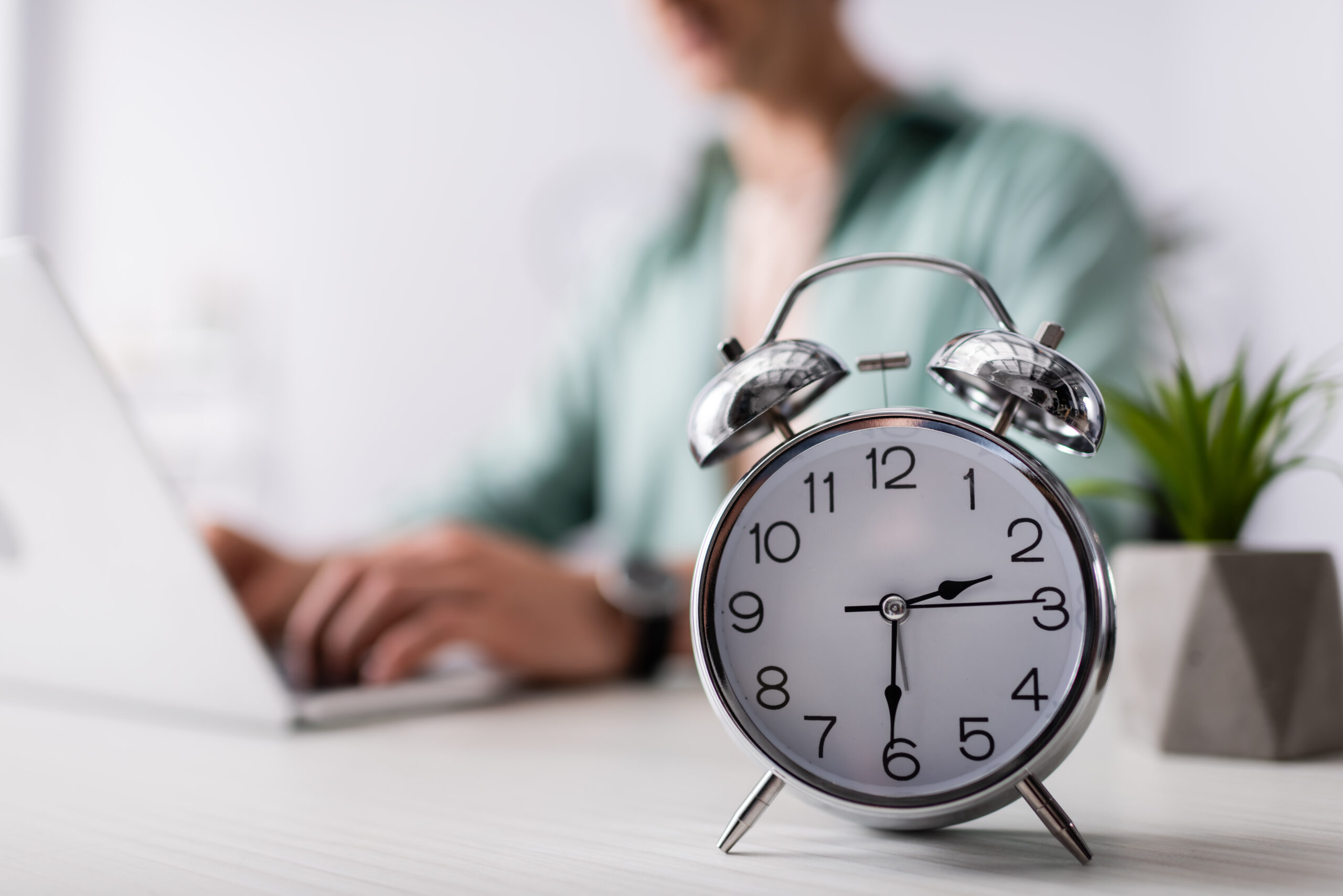 Person working on their laptop at a desk. Clock ticking at the forefront of the image.